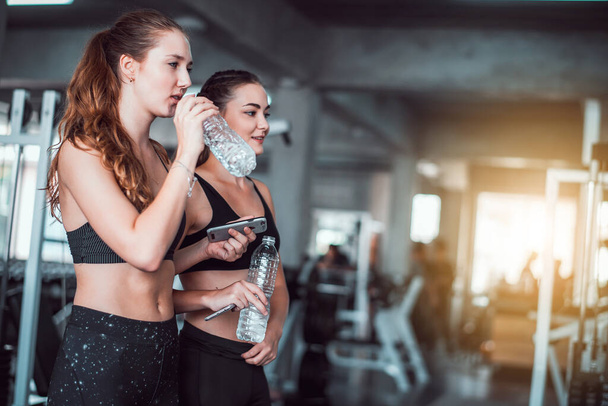 Dwie młode, atrakcyjne, kaukaskie kobiety rozmawiające ze sobą i korzystające ze smartfona lub telefonu komórkowego podczas picia wody i relaksu po ciężkim treningu na siłowni  - Zdjęcie, obraz