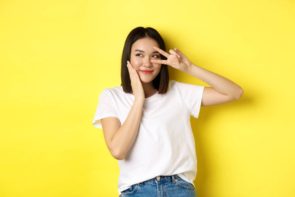 Lovely asian girl in white t-shirt posing with hand on cheek, showing peace sign on eye, standing over yellow background - Foto, Bild