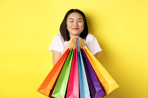 feliz asiático hembra shopper sonriendo y sosteniendo colorido compras bolsas, de pie sobre amarillo fondo - Foto, Imagen