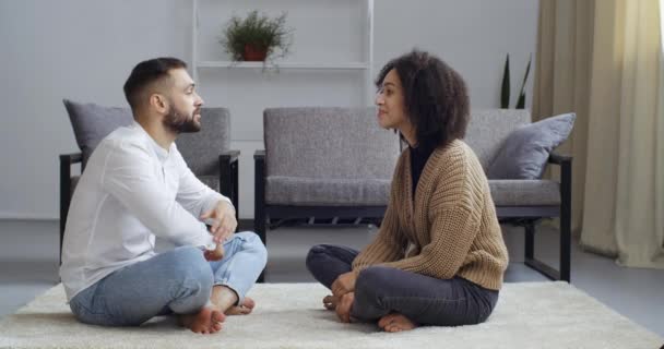Rotujenvälinen pari afrikkalainen amerikkalainen vaimo ja valkoihoinen aviomies puhuvat lattialla lootusasennossa keskustelemassa hymyillen, kaveri siivoaa roskia tytöistä huppari, rakkauden hoito käsite - Materiaali, video