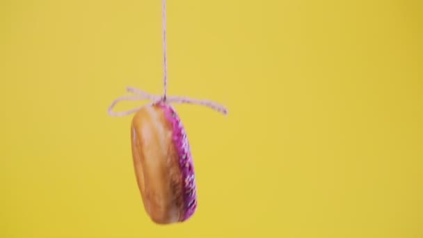 A donut with pink frosting, suspended on a string, and jumps up and down. Yellow background. Soft focus. Close-up. - Footage, Video
