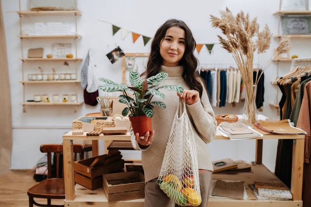 Portrait of a woman in eco shop posing for a photo with net bag and potted plant - 写真・画像