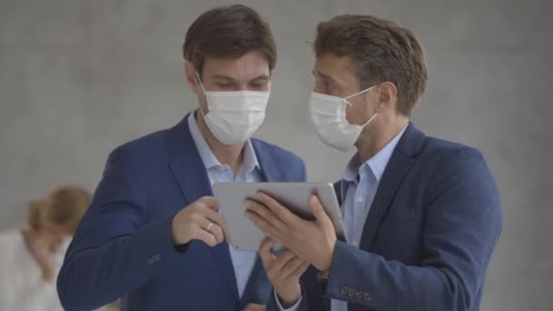 Due bei giovani uomini d'affari con maschere protettive facciali che discutono con tablet digitale in ufficio - Filmati, video