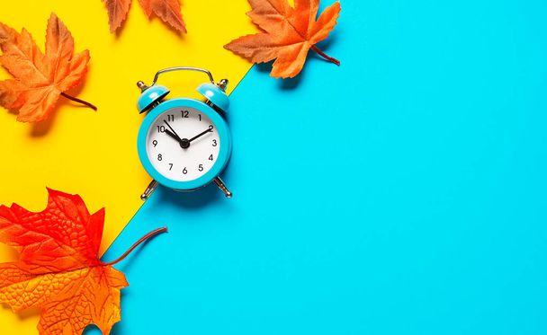 Clock on two tone color yellow and light blue background decorated with colorful fallen leaves. Autumn time concept. Copy space, flat lay - Valokuva, kuva
