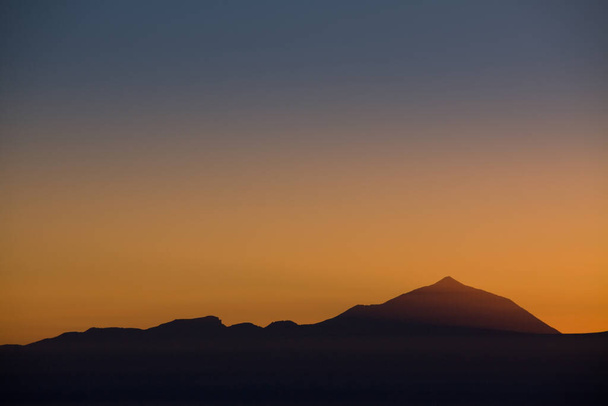 Silhouet van de vulkaan Teide verlicht bij zonsondergang gezien vanaf Gran Canaria in warme oranje tinten - Foto, afbeelding