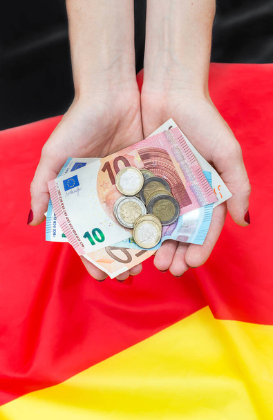 ドイツ国旗の上にユーロ紙幣と硬貨を持つ女性の手. - 写真・画像