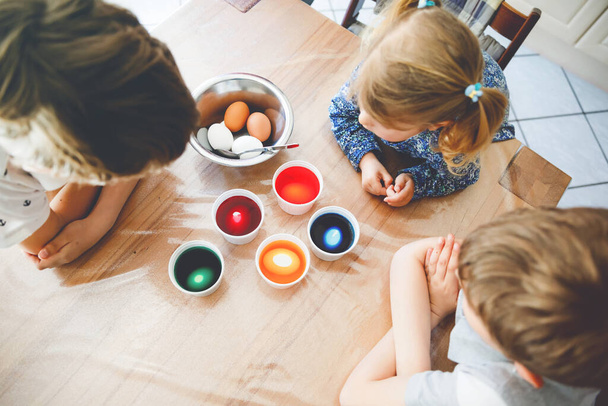 Excited little toddler girl and two kids boys coloring eggs for Easter. Three children, siblings looking surprised at colorful eggs, celebrating holiday with family. From above, unrecognized faces - Photo, Image