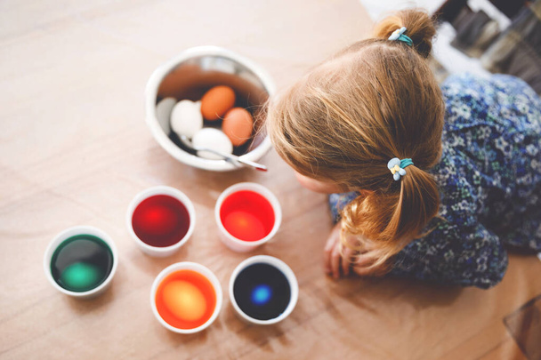 Excited little toddler girl coloring eggs for Easter. Cute happy child looking surprised at colorful colored eggs, celebrating holiday with family. From above, unrecognized face. - Photo, Image