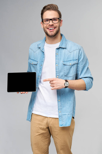 Happy young man wearing jeans shirt standing and holding tablet pc pad over studio grey background - Foto, Imagem