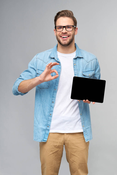 Happy young man wearing jeans shirt standing and holding tablet pc pad over studio grey background - Фото, зображення