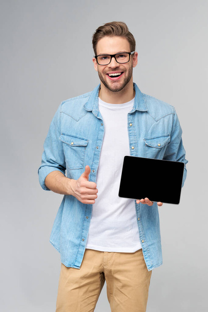 Happy young man wearing jeans shirt standing and holding tablet pc pad over studio grey background - Foto, afbeelding