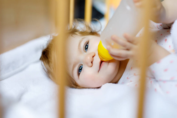 Χαριτωμένο κοριτσάκι κρατώντας μπουκάλι με φόρμουλα ήπια και πόσιμο. Παιδί σε βρεφικό κρεβάτι πριν κοιμηθεί - Φωτογραφία, εικόνα