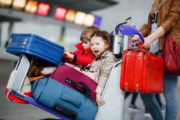 Two little kids, boy and girl, siblings and mother at the airport. Children, family traveling, going on vacation by plane and waiting on trolley with suitcases pushing by woman at terminal for flight. - Zdjęcie, obraz
