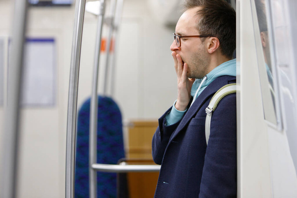 Uomo stanco con gli occhiali nei trasporti pubblici. Giovane maschio esausto in piedi e sbadigliando in metropolitana, assonnato dopo il lavoro va a casa, sfondo sfocato. Affaticamento, concetto di privazione del sonno - Foto, immagini