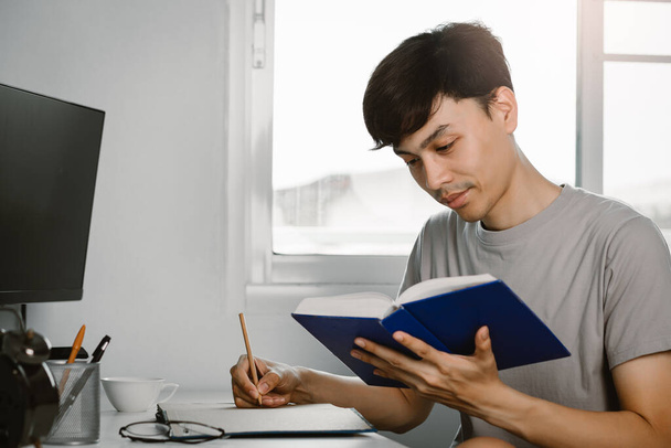 Mladý pohledný asijský muž čtení knihy a psaní v poznámkách na pracovním stole ve volném čase z práce doma, Znalosti a učení koncepce. - Fotografie, Obrázek
