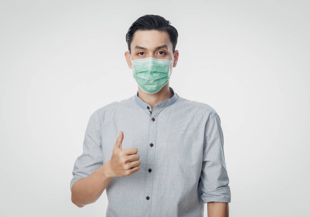 Young Asian Businessman wearing hygienic mask and showing thumb up to prevent infection, 2019-nCoV or coronavirus. Airborne respiratory illness such as pm 2.5 fighting and flu isolated on white background. - Фото, изображение