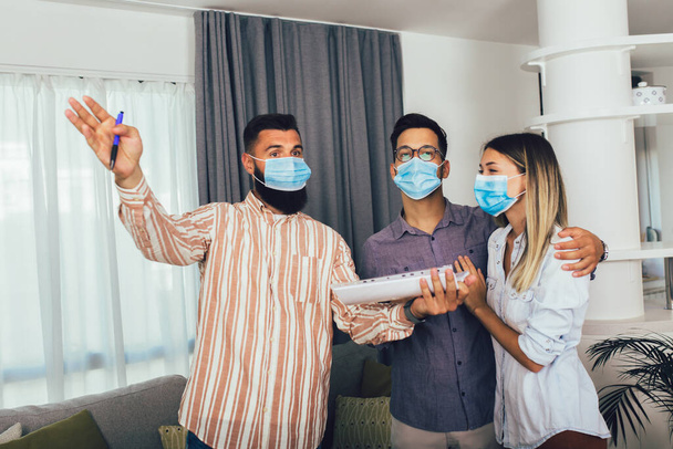 Mladý šťastný manželský pár v lékařských maskách, stojící v novém domě s realitním makléřem. Pandemický koncept. - Fotografie, Obrázek