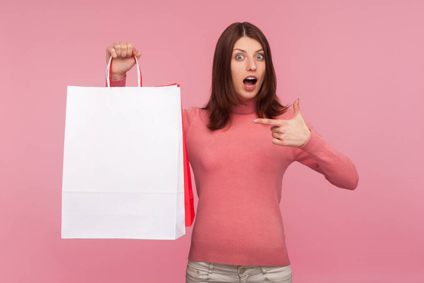 Shocked amazed brunette woman in pink sweater pointing finger at paper bags in her hand, surprised with shopping, low prices good quality. Indoor studio shot isolated on pink background - 写真・画像