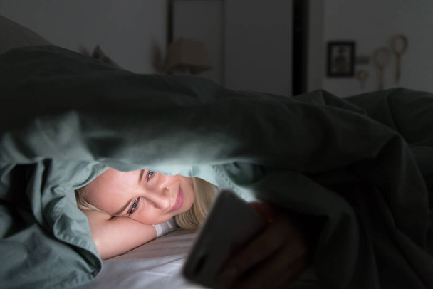 Χαρούμενη νεαρή γυναίκα ξαπλωμένη στο κρεβάτι κάτω από την κουβέρτα χρησιμοποιώντας το κινητό τηλέφωνο αργά το βράδυ, δεν μπορεί να κοιμηθεί. Αϋπνία, νομοφοβία, εθισμός στο διαδίκτυο - Φωτογραφία, εικόνα