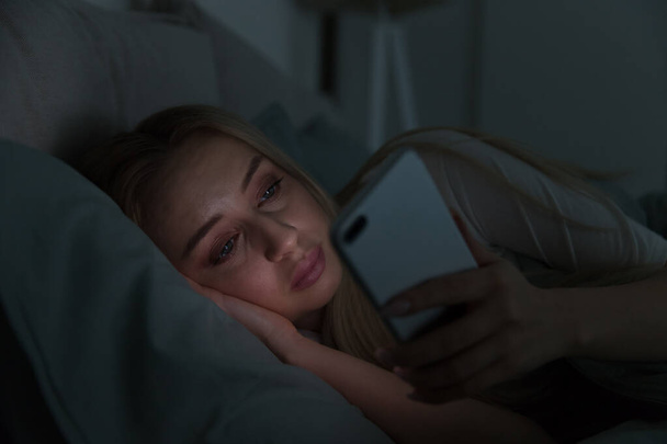 Portrait of young sleepy tired woman lying in bed under the blanket using smartphone at late night, can not sleep/ Insomnia, nomophobia, sleep disorder concept/ Dependency on a cell phone - Φωτογραφία, εικόνα