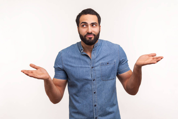Puzzled clueless man with beard in blue shirt throwing up his hands looking at camera with uncertain expression, has no answers. Indoor studio shot isolated on white background - Φωτογραφία, εικόνα