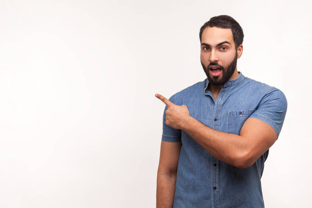 Excited bearded man in blue shirt pointing finger away at empty space, shocked surprised with advertisement area, recommending freespace. Indoor studio shot isolated on white background - Φωτογραφία, εικόνα