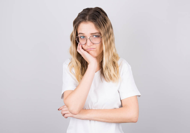 Studio shot of an annoyed annoyed pretty girl in black glasses touching her face and bending over, peeking out from under her eyes, hating talking to a boring person, standing against a gray background - Fotografie, Obrázek