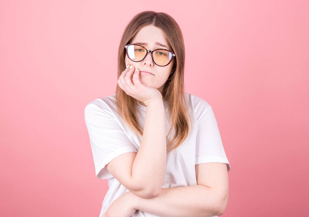 Studio shot of an annoyed annoyed pretty girl in black glasses touching her face and bending over, peeking out from under her eyes, hating talking to a boring person, standing against a pink background - Фото, зображення