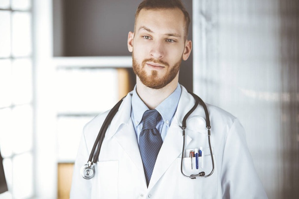 Un docteur à barbe rouge se tenant droit à la clinique près de son lieu de travail. Portrait de médecin. Médecine et concept de santé - Photo, image