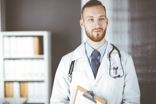 Рыжебородатый врач, стоящий прямо в клинике рядом с рабочим местом. Портрет врача. Медицина и здравоохранение - Фото, изображение