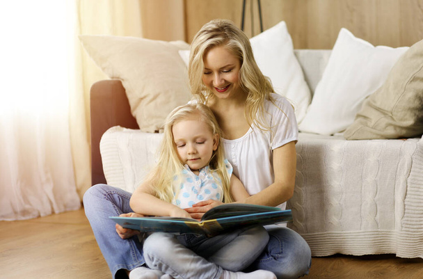 Happy family. Blonde young mother reading a book to her cute daughter while sitting at wooden floor in sunny room. Motherhood concept - Foto, Imagem