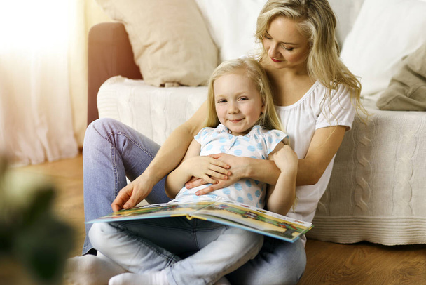 Happy family. Blonde young mother reading a book to her cute daughter while sitting at wooden floor in sunny room. Motherhood concept - Foto, immagini