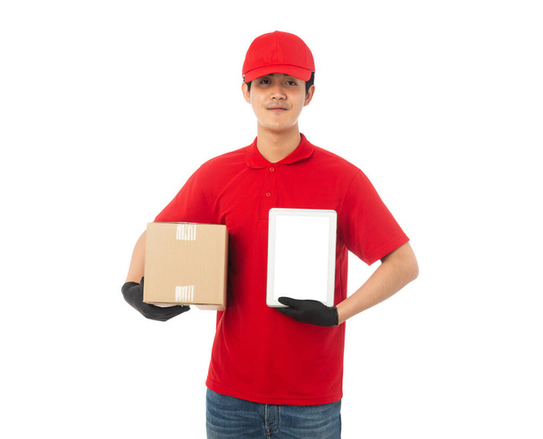 Kırmızı üniformalı genç teslimatçı elinde karton kutu ve tablet bilgisayar modeliyle beyaz arka planda kırpma yolu ile izole edilmiş.. - Fotoğraf, Görsel
