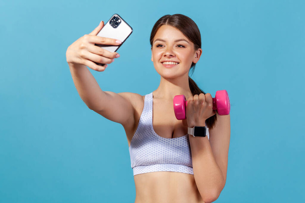 Excited sports woman in white top making selfie exercising with dumbbell, monitoring her indicators on smartwatch through app on smartphone, blogging. Indoor studio shot isolated on blue background - Φωτογραφία, εικόνα