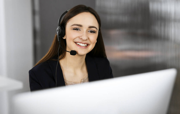 Young smiling girl in headsets is talking to the firms client, while sitting at the desk in a companys office. Portrait of a call center operator at work - Foto, Imagen