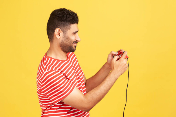 Profile portrait bearded man in red striped t-shirt holding in hands red gamepad joystick, grimacing playing video games. Indoor studio shot isolated on yellow background - Foto, afbeelding