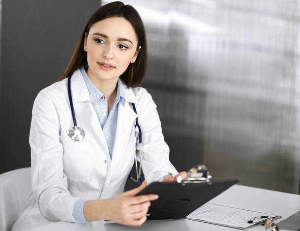 Smiling young woman-doctor is filling up medical recommendations using a clipboard. Therapist at work in a clinic office. Medicine concept - Photo, image