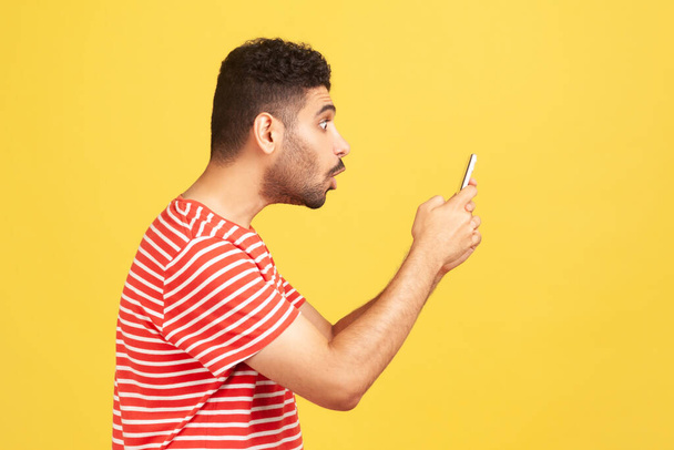 Profile portrait surprised shocked man with beard in red striped t-shirt looking at smartphone display with astonishment, confused with tariffs. Indoor studio shot isolated on yellow background - Zdjęcie, obraz