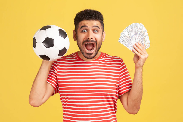 Extremely excited happy man with beard in striped t-shirt holding soccer ball and hundred dollar bills, screaming looking camera, betting and winning. Indoor studio shot isolated on yellow background - Фото, зображення