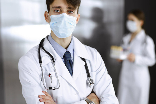 Young male doctor standing with arms crossed in clinic. Smart physician wearing medical protective mask in modern hospital. Medicine and Corona virus concept - Foto, Bild