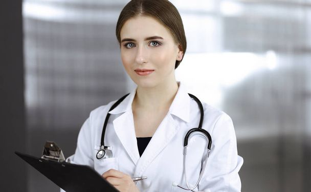 Friendly female doctor standing and holding clipboard in modern clinic. Portrait of cheerful smiling physician - Fotoğraf, Görsel