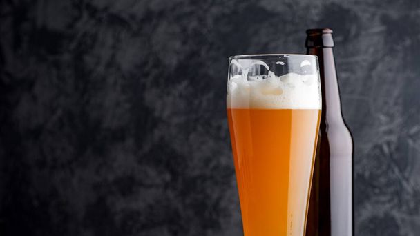 Пшеничное нефильтрованное пиво в стакане ярко насыщенного цвета и пивная бутылка на темном фоне - Фото, изображение