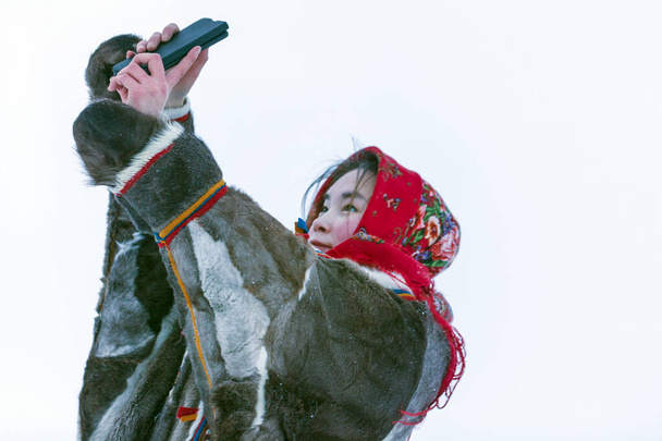 Tundra 'nın kuzeyindeki milli kış kıyafetleri içindeki genç bir kız akıllı telefondan selfie çekiyor. - Fotoğraf, Görsel