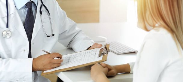 Doctor and patient discussing medical exam results while sitting at the desk in sunny clinic, close-up. Male physician using clipboard. - Foto, Imagen