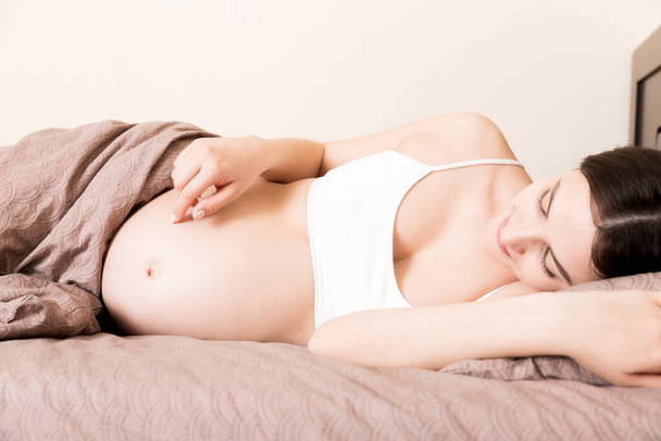 terhes nő ül az ágyban, és megérinti a hasát otthon. terhességi várakozási koncepció. - Fotó, kép