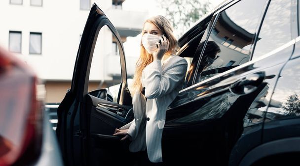 Eine junge Geschäftsfrau in Schutzmaske telefoniert und fährt bei einer Pandemie Auto  - Foto, Bild