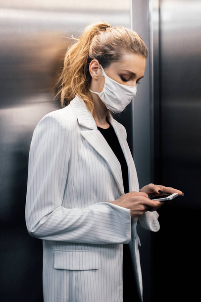 Молодий працівник, бізнес-леді, носить маску безпеки і використовує телефон в ліфті
  - Фото, зображення
