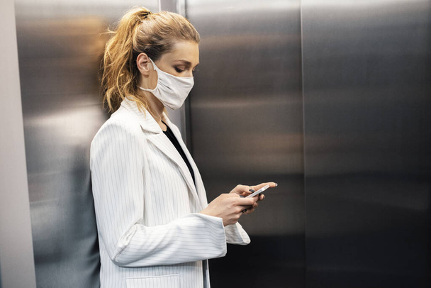 Mladá pracovnice, obchodnice, nosí bezpečnostní masku a používá telefon ve výtahu.  - Fotografie, Obrázek