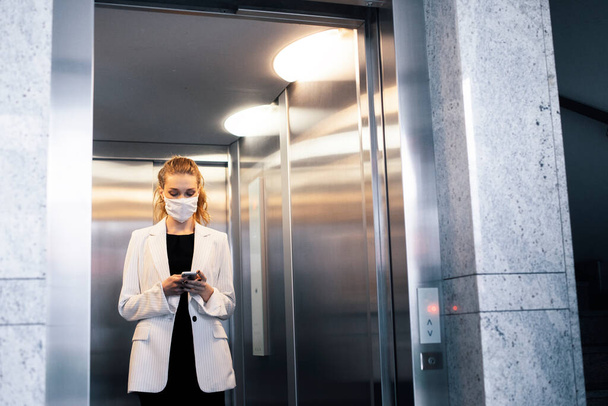 Eine junge Arbeiterin, Geschäftsfrau trägt eine Schutzmaske und telefoniert im Aufzug  - Foto, Bild