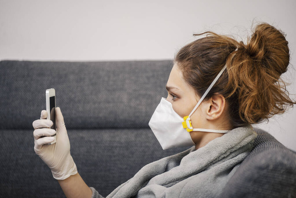 A young woman in a protective mask and gloves is using the phone during the quarantine - 写真・画像